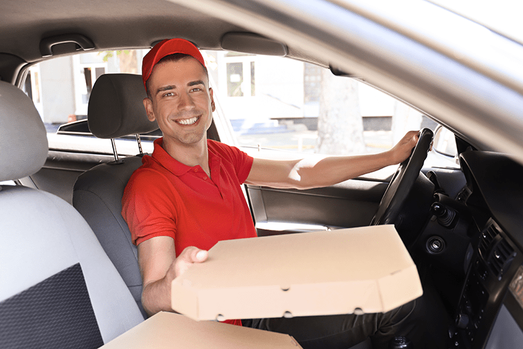Smiling male sitting behind the wheel of his car. He is holding out a cardboard box with food towards the passenger window.
