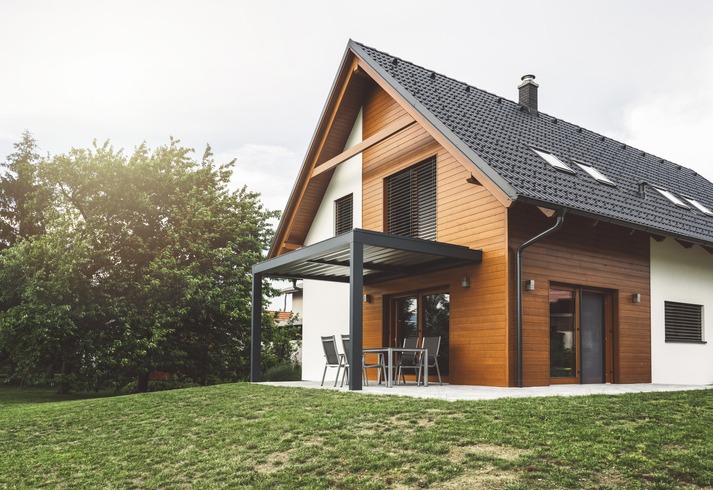 Exterior picture of a eco-friendly, minimalist vacation home. The siding is a combination of wood and concrete with a black slate roof and deck cover. There is a patio table set and a gently sloping lawn with trees in the background.