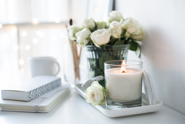 White room interior decor with burning hand-made candle and bouquet of fresh roses on table, luxury home decorations in daylight closeup.
